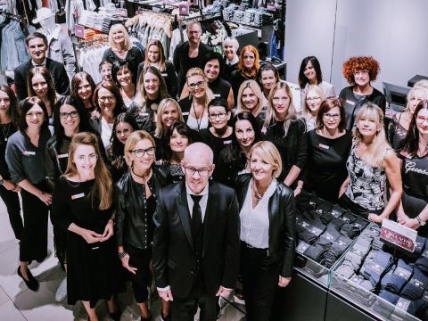 Eine Gruppe von Menschen, in formeller und legerer Kleidung, steht zusammen in einem Bekleidungsgeschäft. Sie lächeln und sind von Kleiderständern umgeben, was auf ein Teamtreffen oder eine Veranstaltung hindeutet.