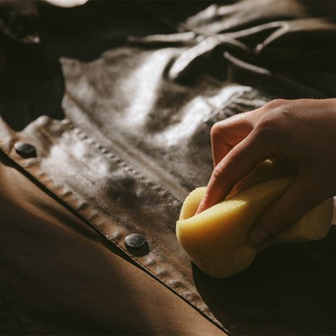Eine Hand reinigt oder poliert mit einem gelben Schwamm eine braune Lederjacke. Dabei konzentriert sie sich auf Knöpfe und Nähte. Die Beleuchtung hebt die Struktur und den Glanz des Leders hervor.
