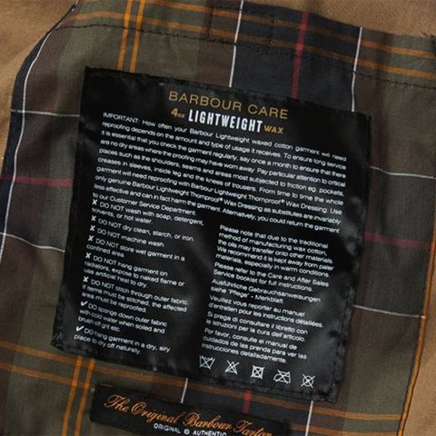 Nahaufnahme eines Pflegeetiketts auf kariertem Stoff. Das Etikett enthält Pflegehinweise für die Lightweight Wax-Kleidung von Barbour sowie Ratschläge zur Reinigung und Pflege. Der Hintergrund zeigt ein braunes, grünes und oranges Schottenmuster.