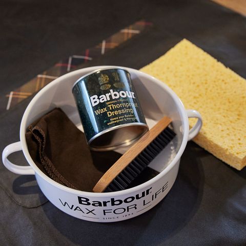 Ein Barbour-Pflegeset mit einer Dose Wachspflege, einem Poliertuch, einer Bürste und einem gelben Schwamm. Die Artikel sind in einem runden weißen Behälter mit dem Text „Barbour Wax for Life“ angeordnet.