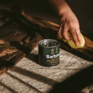 Eine Person trägt mit einem gelben Schwamm Wachs auf ein braunes Kleidungsstück auf. Eine Dose mit der Aufschrift „Barbour Wax Thornproof Dressing“ steht in der Nähe auf einer mit Stoff bedeckten Oberfläche. Die Szene ist warm beleuchtet, und die Schatten lassen auf eine natürliche Lichtquelle schließen.