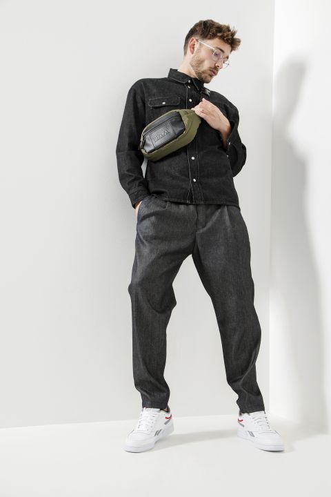 Eine Person in schwarzer Jeansjacke, grauer Hose und weißen Turnschuhen posiert vor einer weißen Wand. Sie trägt eine Brille und eine grüne Umhängetasche mit einer schwarzen Vordertasche.