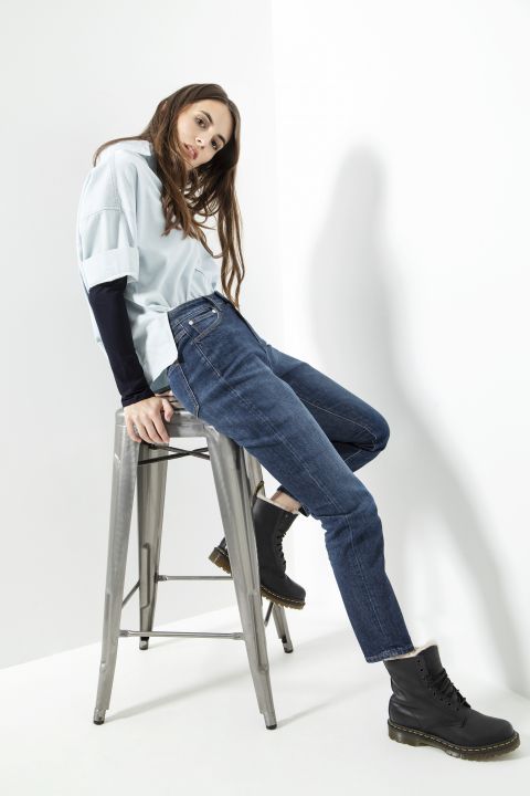 Eine Frau in hellblauem Top und dunkler Jeans sitzt lässig auf einem silbernen Hocker vor weißem Hintergrund. Sie trägt schwarze Stiefel und lehnt sich leicht nach hinten, wodurch sie eine entspannte und stilvolle Pose einnimmt.