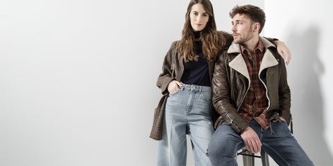 Eine Frau in kariertem Mantel und Jeans steht selbstbewusst neben einem Mann, der auf einem Hocker sitzt und eine braune Lederjacke und Jeans trägt. Beide sehen in lässigen Outfits vor einem schlichten weißen Hintergrund stilvoll und entspannt aus.