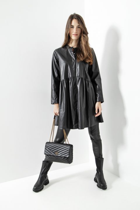 Eine Frau in einer langen schwarzen Lederjacke und schwarzen Lederhosen posiert selbstbewusst. Sie hält eine schwarze gesteppte Handtasche mit Kettenriemen und trägt schwarze Stiefel. Die Kulisse ist eine minimalistische weiße Ecke.