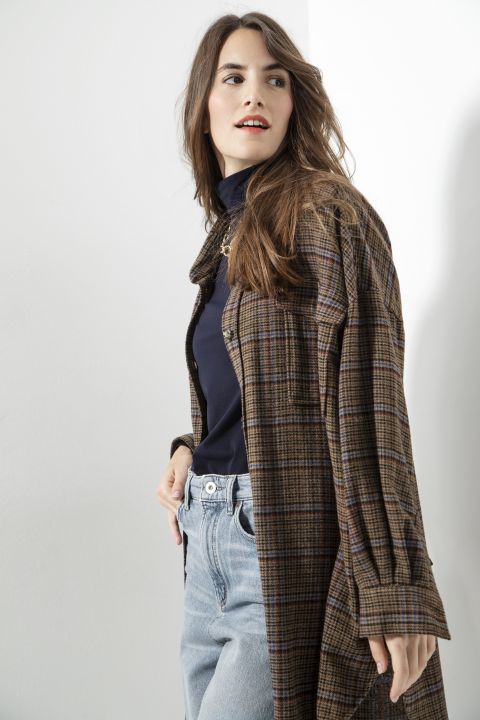 Eine Person mit langen Haaren trägt einen braun karierten Mantel über einem schwarzen Rollkragenpullover und hellblaue Jeans und blickt zur Seite. Im Hintergrund ist eine schlichte, helle Wand zu sehen.