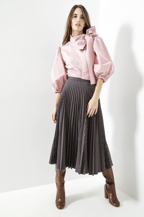 Eine Frau posiert elegant in einer hellrosa Bluse mit Puffärmeln und einer großen Schleife. Dazu trägt sie einen plissierten, karierten Rock und kniehohe braune Stiefel. Sie steht vor einer weißen Wand und wirft einen Schatten leicht nach links.