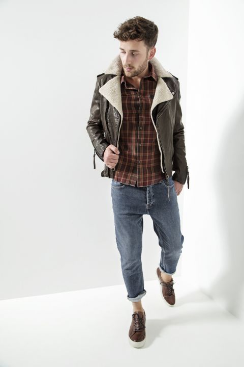Ein Mann mit Bart trägt eine braune Lederjacke mit Lammfellkragen über einem karierten Hemd. Dazu trägt er blaue Jeans und braune Schuhe. Er blickt nach links, während er in einer weißen, minimalistischen Umgebung steht.