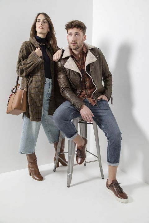 Eine Frau in kariertem Mantel und Jeans steht neben einem sitzenden Mann in Lederjacke und hochgekrempelten Jeans. Beide tragen lässige, elegante Kleidung und braune Schuhe und posieren vor einem hellen Hintergrund.