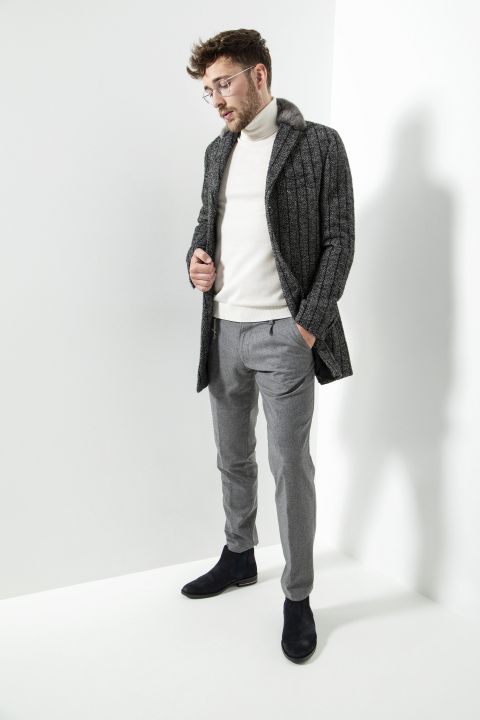Ein Mann mit Brille posiert in einem stylischen Outfit, bestehend aus einem strukturierten grauen Mantel, einem weißen Rollkragenpullover, grauen Hosen und schwarzen Schuhen. Er steht vor einem minimalistischen weißen Hintergrund, blickt nach unten und hat eine Hand in der Manteltasche.