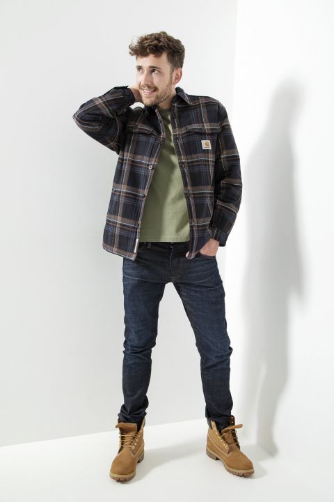 Ein Mann posiert vor einer hellgrauen Wand. Er trägt ein kariertes Hemd über einem grünen T-Shirt, dunkle Jeans und hellbraune Stiefel. Er hat eine entspannte Haltung, eine Hand im Nacken, lächelt und wirft einen weichen Schatten auf die Wand.