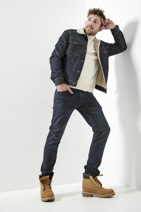 Eine Person posiert selbstbewusst in einem stylischen Outfit bestehend aus einer Jeansjacke mit Lammfellkragen, einem cremefarbenen Pullover, dunklen Jeans und hellbraunen Stiefeln. Die Kulisse ist minimalistisch mit weißem Hintergrund.
