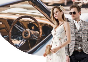 Eine Frau im weißen Kleid und ein Mann im karierten Anzug stehen selbstbewusst neben einem Bild des hölzernen Lenkrads und Armaturenbretts eines Oldtimers. Die Szene vereint Eleganz und klassischen Automobilstil.