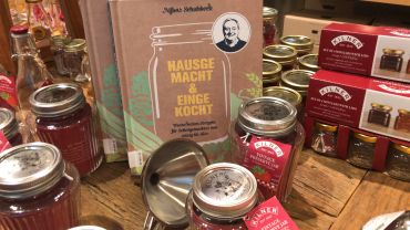 Eine Ausstellung von Kochbüchern, darunter „Hause Macht & Enge Kocht“ mit einer Glasillustration und einem Porträt, umgeben von Kilner-Gläsern mit roter Konfitüre. Die Artikel sind auf einem Holztisch angeordnet.