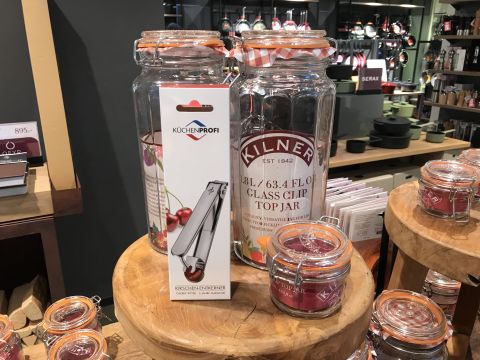 Ein Schaufenster mit großen Bügelgläsern von Kilner und kleineren Varianten neben einer Schachtel für einen Glasöffner von Küchenprofi. Die Gläser sind durchsichtig und haben rot karierte Deckel. Im Hintergrund sind Kochutensilien und Produkte zu sehen.