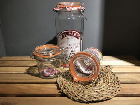 Drei Kilner-Gläser mit orangefarbenen Siegeln auf einer Holzoberfläche. Der größte Behälter steht aufrecht und trägt die Aufschrift „1,8 l/63,4 fl oz Glas mit Clipverschluss“. Die kleineren Behälter stehen neben und auf einer geflochtenen Matte, einer liegt auf der Seite.