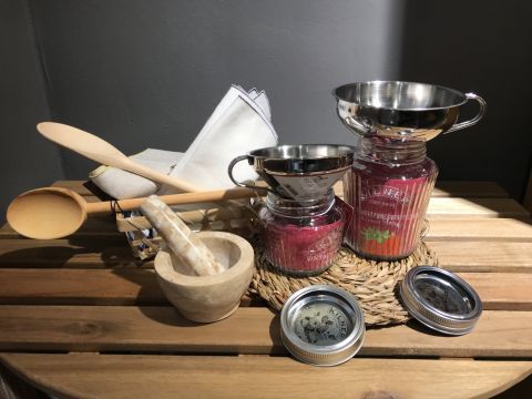 Kücheneinrichtung mit Holzlöffeln, einem Mörser und Stößel, Glasgefäßen mit Metalldeckeln und einem weißen Tuch. Die Gefäße sind mit rosa und rotem Inhalt gefüllt. Sie stehen auf einer Holzoberfläche und werden durch natürliches Licht hervorgehoben.