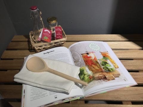 Ein aufgeschlagenes Kochbuch mit Abbildungen von eingelegtem Gemüse in Gläsern. Daneben liegt ein Holzlöffel auf einer weißen Serviette. Im Hintergrund ist ein kleiner Korb mit einer Glasflasche und zwei Gewürzpaketen zu sehen.