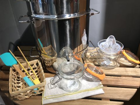 Ein großer Suppentopf aus Edelstahl steht auf einer Holzoberfläche. Daneben stehen zwei Gläser mit orangefarbenen Deckeln, eine Zitruspresse aus Glas, eine Serviette und ein Korb mit einem Spatel und Holzutensilien.