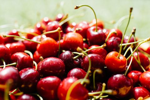Eine Nahaufnahme eines Stapels reifer roter Kirschen mit grünen Stielen, die im Sonnenlicht baden. Die Kirschen haben einen glänzenden Schimmer und der Hintergrund ist leicht verschwommen, was die leuchtende Farbe und Frische der Frucht betont.