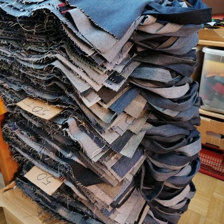 Auf einem Karton liegt ein großer Stapel ordentlich aufgestapelter Jeansstoffstücke, bereit zum Zusammensetzen. Die Stoffkanten sind leicht ausgefranst, und aus den Stapeln lugen Etiketten mit Nummern hervor. Der Raum hat einen Holzboden.