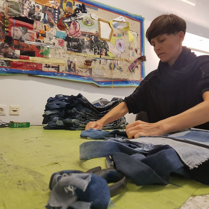 Eine Person mit kurzen braunen Haaren arbeitet an einem Tisch und schneidet Jeansstoffstücke zu. Der Arbeitsplatz hat bunte Collagen an den Wänden und eine grüne Oberfläche voller Stoffe und Scheren. Im Hintergrund ist eine Steckdosenleiste zu sehen.