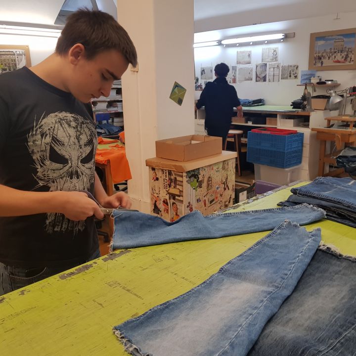 Ein junger Mensch im schwarzen T-Shirt schneidet Jeansstoff auf einem großen Arbeitstisch zu. Im Hintergrund arbeiten weitere Menschen in einer Werkstatt voller Nähmaschinen und verschiedener Materialien.