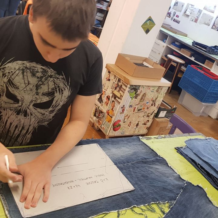 Eine Person arbeitet in einer Werkstatt an einem Denim-Projekt und zeichnet ein Muster auf Stoff. Der Arbeitsbereich ist mit verschiedenen Textilien, Kisten und Wanddekorationen gefüllt, was eine kreative und fleißige Atmosphäre schafft.