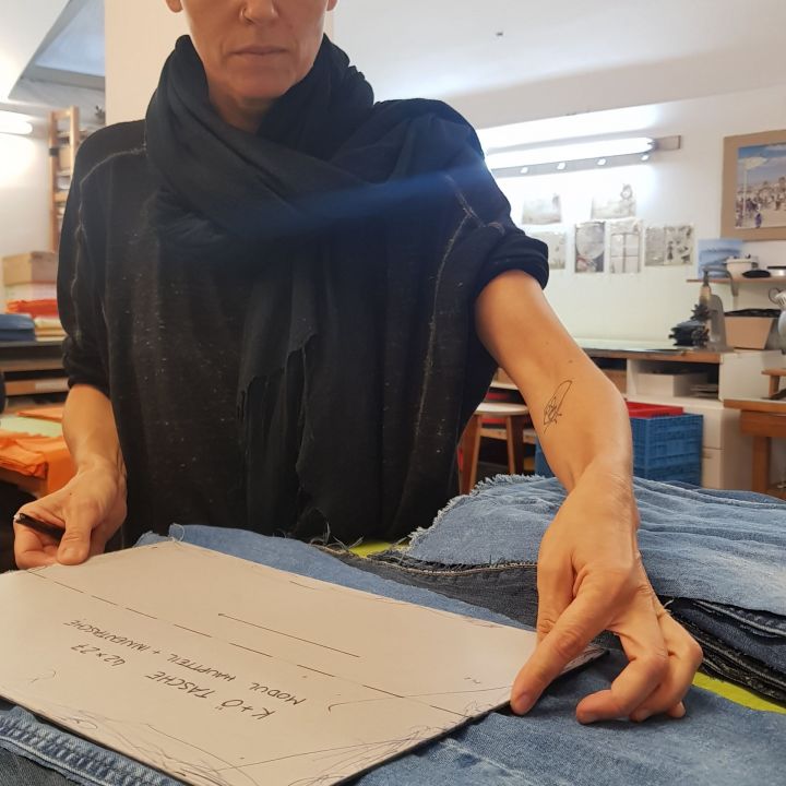 Eine Person im dunklen Hemd arbeitet in einer Werkstatt mit einem Papierschnittmuster auf blauem Jeansstoff. Im Hintergrund sind Regale mit Materialien und ein beleuchteter Arbeitsplatz zu sehen.