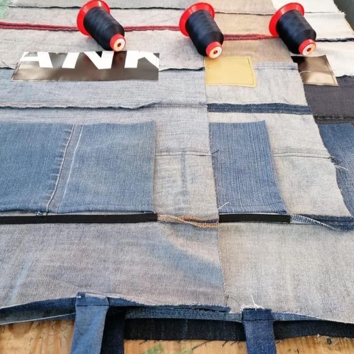 Ein Patchwork-Stofflayout auf einem Tisch mit verschiedenen Jeansstücken, die in einem Gittermuster zusammengenäht sind. Drei Spulen dunkelblauen Garns sind auf dem Stoff aufgereiht. In der oberen linken Ecke ist ein kleines weißes Etikett zu sehen.