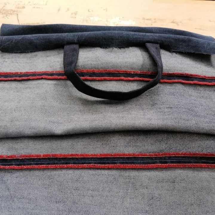 Teilweise genähter Jeansstoff mit roten Nähten liegt auf einer flachen Oberfläche und weist einen Schlaufengriff auf, der an die frühen Phasen der Herstellung einer Tasche oder eines Kleidungsstücks erinnert.