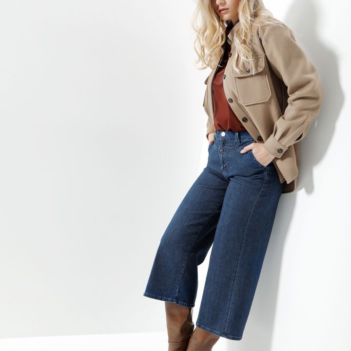 Eine Person mit langen blonden Haaren lehnt an einer weißen Wand. Sie trägt eine hellbraune Jacke über einem braunen Oberteil, blaue Jeans mit weitem Bein und kniehohe braune Stiefel. Die Umgebung ist gut beleuchtet, wodurch die Texturen und Farben des Outfits hervorgehoben werden.