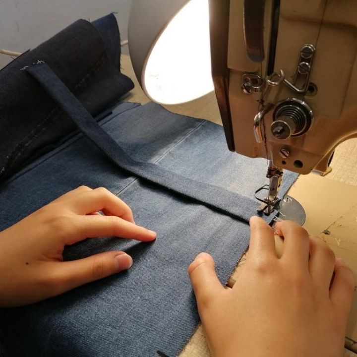 Hände bedienen eine Nähmaschine und nähen blauen Jeansstoff. Eine Lampe beleuchtet den Arbeitsbereich und hebt die Hände und das genähte Material hervor.