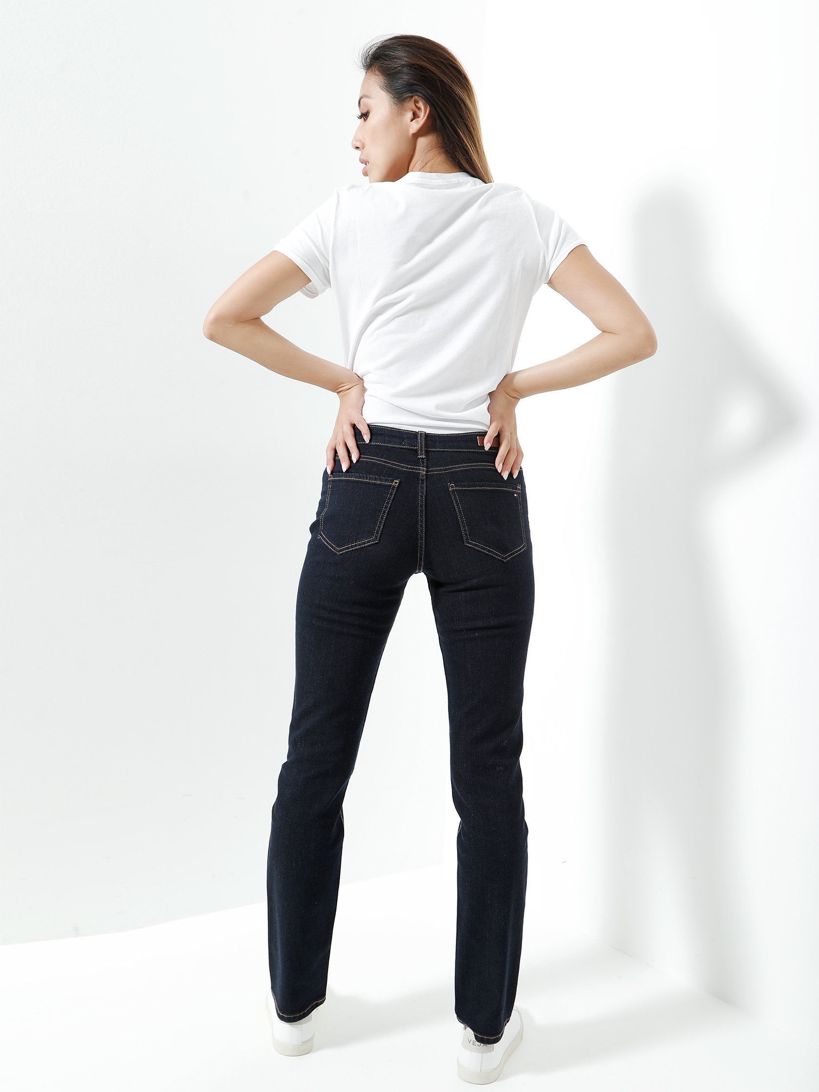 Eine Person steht vor einem weißen Hintergrund und blickt von der Kamera weg. Sie trägt ein weißes T-Shirt, dunkle Jeans und weiße Turnschuhe und hat die Hände in die Hüften gestemmt.