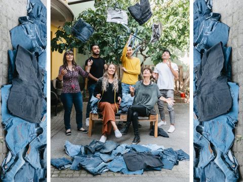 Eine Gruppe von sechs Personen steht im Freien, umgeben von Blue Jeans. Drei sitzende Personen halten Jeans, während die anderen sie in die Luft werfen, wodurch eine lebendige, verspielte Szene entsteht. Ein belaubter Baum im Hintergrund fügt ein natürliches Element hinzu.