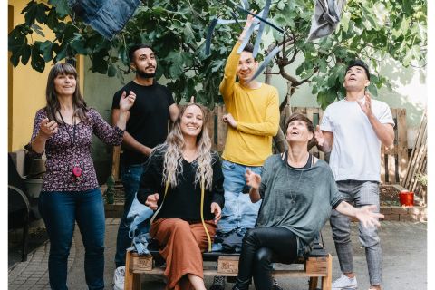 Eine Gruppe von sechs Personen steht und sitzt unter einem großen Baum. Sie lächeln und einige werfen spielerisch ihre Jacken in die Luft. Die Szene ist lebhaft und fröhlich, das Sonnenlicht fällt durch die Blätter.