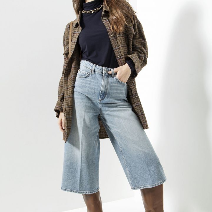 Eine Frau mit langen Haaren lächelt beim Posieren. Sie trägt eine braun karierte Jacke über einem schwarzen Oberteil, weite Bluejeans und braune kniehohe Stiefel. Sie steht vor einem schlichten, hellen Hintergrund.