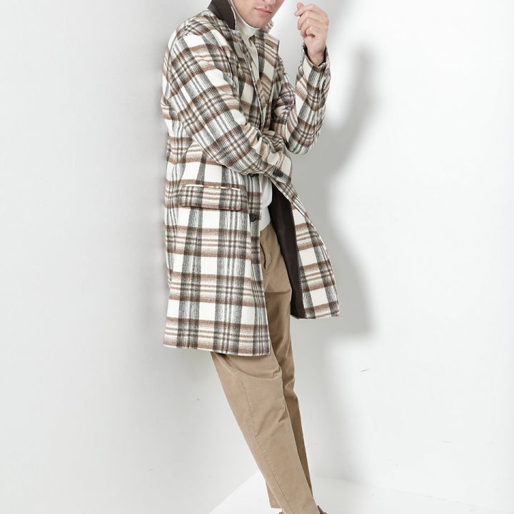 Ein Mann in kariertem Mantel, beiger Hose und braunen Schuhen posiert vor einer weißen Wand. Er lehnt sich leicht nach vorne, eine Hand liegt nahe am Kinn, und blickt in die Kamera. Das Outfit ist lässig und stylisch.
