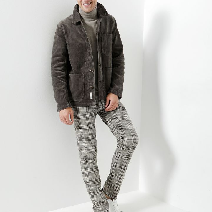Eine Person trägt eine dunkle Jacke über einem Rollkragenpullover und karierten Hosen und lehnt lächelnd an einer weißen Wand. Weiße Sneakers runden das Outfit ab. Das minimalistische Ambiente verleiht dem Ganzen einen modernen Touch.