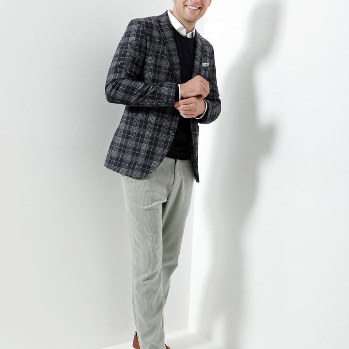Ein Mann steht lächelnd da und trägt einen karierten Blazer, einen schwarzen Pullover, ein weißes Hemd, hellgraue Hosen und braune Schuhe. Er ist leicht zur Seite gedreht, hat einen fröhlichen Gesichtsausdruck und posiert vor einem schlichten weißen Hintergrund.