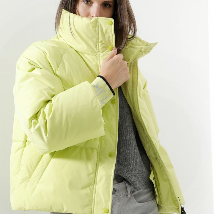 Eine Person trägt eine hellgelbe Pufferjacke, deren Kragen ihr Gesicht teilweise bedeckt. Sie hat langes braunes Haar und trägt einen grauen Pullover und hellgraue Hosen. Der Hintergrund ist in einer schlichten hellen Farbe gehalten.