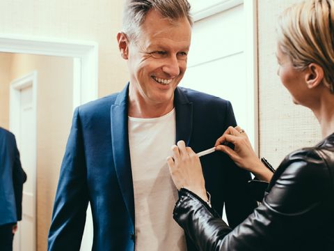 Eine Person bekommt von einem Schneider einen Anzug angepasst. Sie trägt ein weißes Hemd und einen blauen Blazer. Der Schneider, der eine schwarze Jacke trägt, passt den Blazer an, während die Person lächelt. Die Szene scheint ein Bekleidungsgeschäft oder eine Umkleidekabine zu sein.