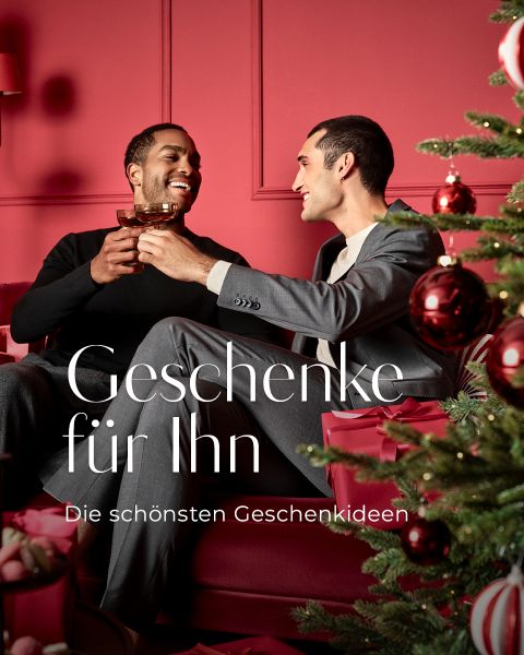 Zwei Männer feiern mit Getränken in einem festlichen, roten Weihnachtszimmer neben einem geschmückten Weihnachtsbaum. Text: Geschenke für Ihn, Die schönsten Geschenkideen.