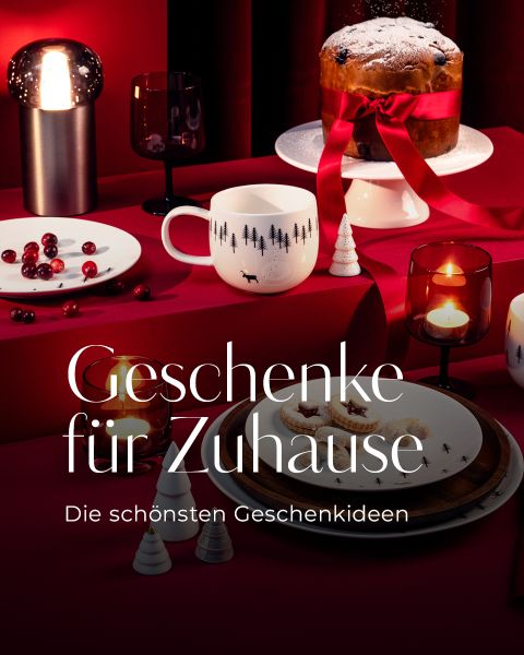 Ein festlicher Tisch mit Kerzen, Tassen, Keksen, Preiselbeeren und einem mit Bändern umwickelten Kuchen. Der Text lautet: "Geschenke für Zuhause Die schönsten Geschenkideen.