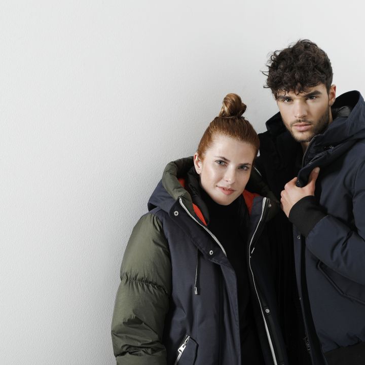 Eine Person mit hochgesteckten langen Haaren trägt eine olivgrüne und schwarze Jacke. Daneben trägt eine Person mit lockigem Haar eine dunkelblaue Jacke. Beide stehen vor einem weißen Hintergrund und blicken in die Kamera.