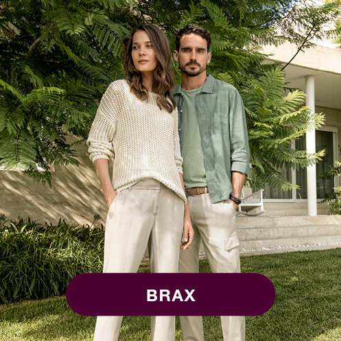 Eine Frau in einem cremefarbenen Pullover und beigen Hosen steht neben einem Mann in einem grünen Hemd und beigen Hosen in einem Garten mit üppigem Grün. Sie erscheinen vor einem modernen Gebäude. Das Wort „BRAX“ steht auf einem violetten Rechteck am unteren Rand.