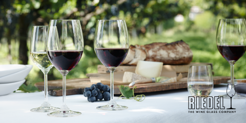 Im Freien steht ein Tisch mit vier Weingläsern, gefüllt mit Rot- und Weißwein. Im Hintergrund sind Brot, Käse und Trauben zu sehen, mit üppigem Grün. In der unteren rechten Ecke ist das Logo „Riedel: The Wine Glass Company“ zu sehen.