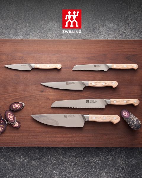 Fünf Küchenmesser mit hellen Holzgriffen liegen auf einer Holzunterlage, daneben liegen in Scheiben geschnittene violette Möhren, und darüber ist das Zwilling-Logo zu sehen.