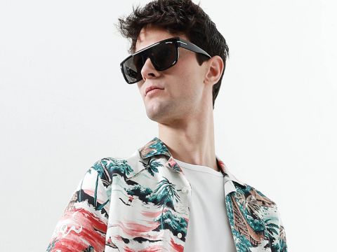 Eine Person mit einer großen schwarzen Sonnenbrille und einem bunten Hemd mit Tropenmuster blickt vor einem schlichten Hintergrund zur Seite.