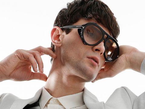 Eine Person mit kurzen dunklen Haaren trägt eine übergroße Sonnenbrille und einen hellen Anzug. Sie hat ihre Hände nahe am Hals positioniert und ihren Kopf leicht nach hinten geneigt. Der Hintergrund ist schlicht weiß.